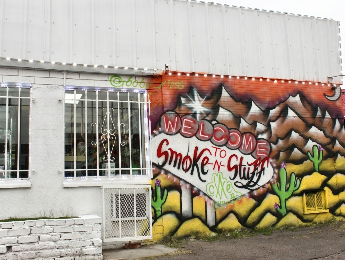 Hand-painted desert-marquee mural reading 'Welcome to Smoke-n-Stuff' on the shop's barred-window storefront.